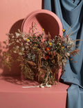 A dried floral arrangement in a terracotta-colored vase, styled on a matching terracotta pedestal with a draped blue fabric backdrop and a large oval mirror behind it.