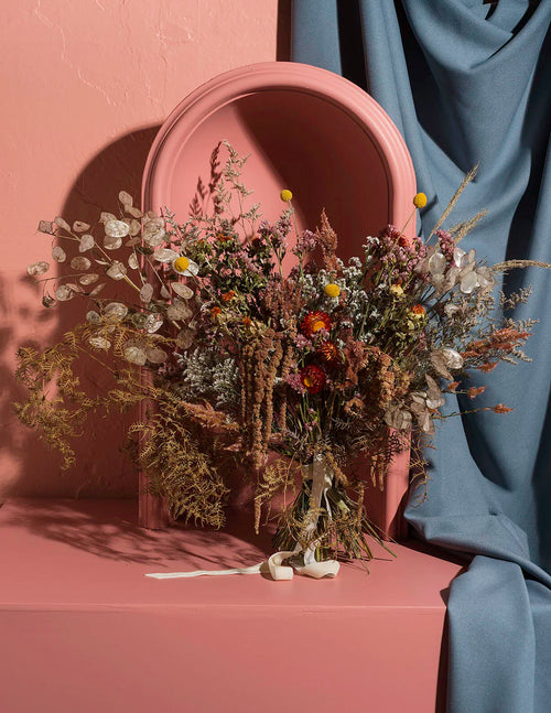 A dried floral arrangement in a terracotta-colored vase, styled on a matching terracotta pedestal with a draped blue fabric backdrop and a large oval mirror behind it.
