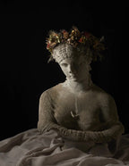 A classical-style stone statue bust of a draped female figure with arms crossed, wearing a floral crown, set against a black background with draped fabric in the foreground.