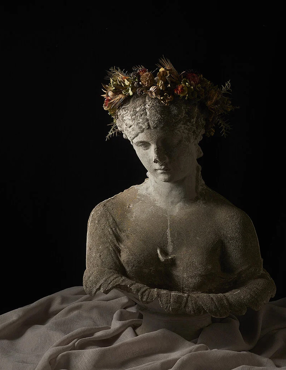 A classical-style stone statue bust of a draped female figure with arms crossed, wearing a floral crown, set against a black background with draped fabric in the foreground.