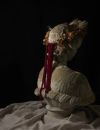 A classical-style stone bust sculpture shown from the back and side, wearing an ornate floral headpiece with dried flowers and long red ribbon tails, set against a black background with draped fabric at the base.