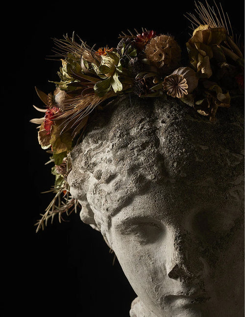 A classical stone bust of a woman's face with a dried floral and botanical crown against a solid black background.