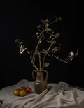 A rustic ceramic vase filled with dried branches and small berries, arranged on draped beige fabric with a few tomatoes in the foreground against a black background.