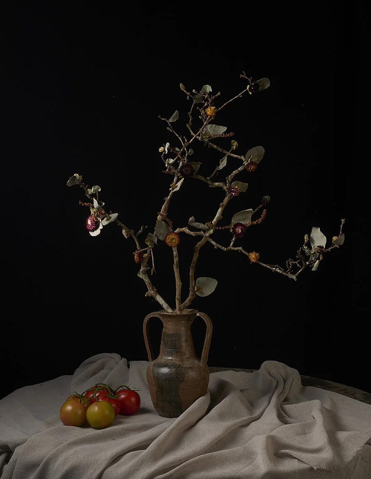 A rustic ceramic vase filled with dried branches and small berries, arranged on draped beige fabric with a few tomatoes in the foreground against a black background.