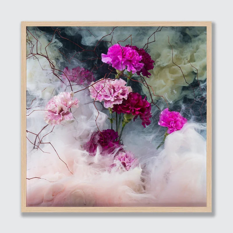 A framed square art print featuring pink and magenta flowers emerging from soft white and pale pink smoke against a dark smoky background, set in a light natural wood frame.