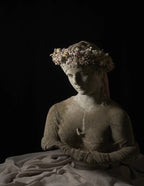 A classical-style stone bust sculpture of a woman wearing a floral crown, draped in pale fabric, set against a solid black background.