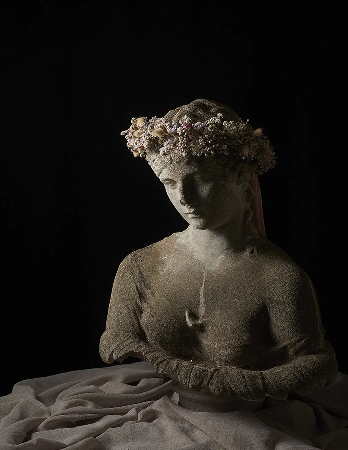 A classical-style stone bust sculpture of a woman wearing a floral crown, draped in pale fabric, set against a solid black background.