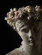 A close-up studio shot of a classical stone bust with a crown of dried flowers and soft botanicals against a solid black background.