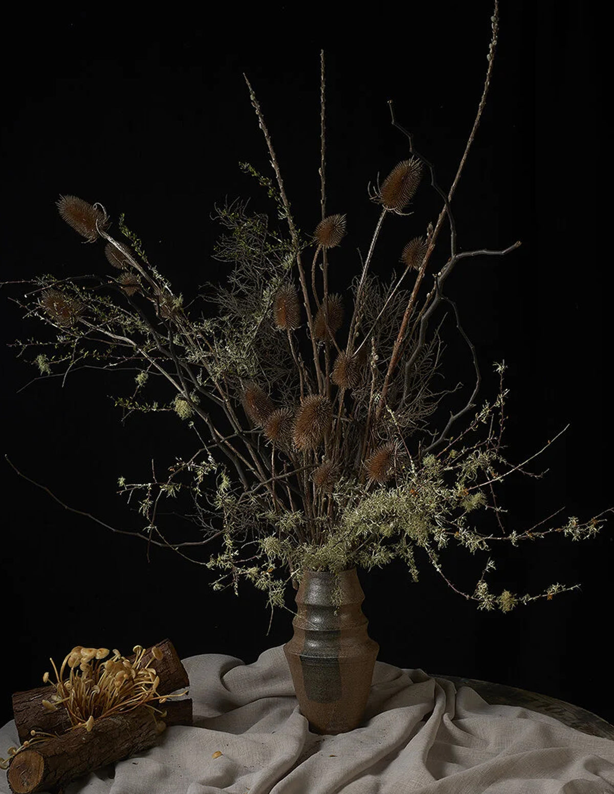 A rustic dried flower arrangement featuring brown seed heads, delicate pale green stems, and branches, presented in a textured brown ceramic vase on draped beige fabric against a solid black background.