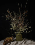 A dried floral arrangement with brown seed pods and pale greenish stems in a textured olive-green ceramic vase, set on draped fabric against a black background with a small log prop at the lower left.