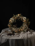 A rustic dried flower wreath with beige, white, and brown botanical elements, displayed on a draped beige fabric-covered table against a solid black background.