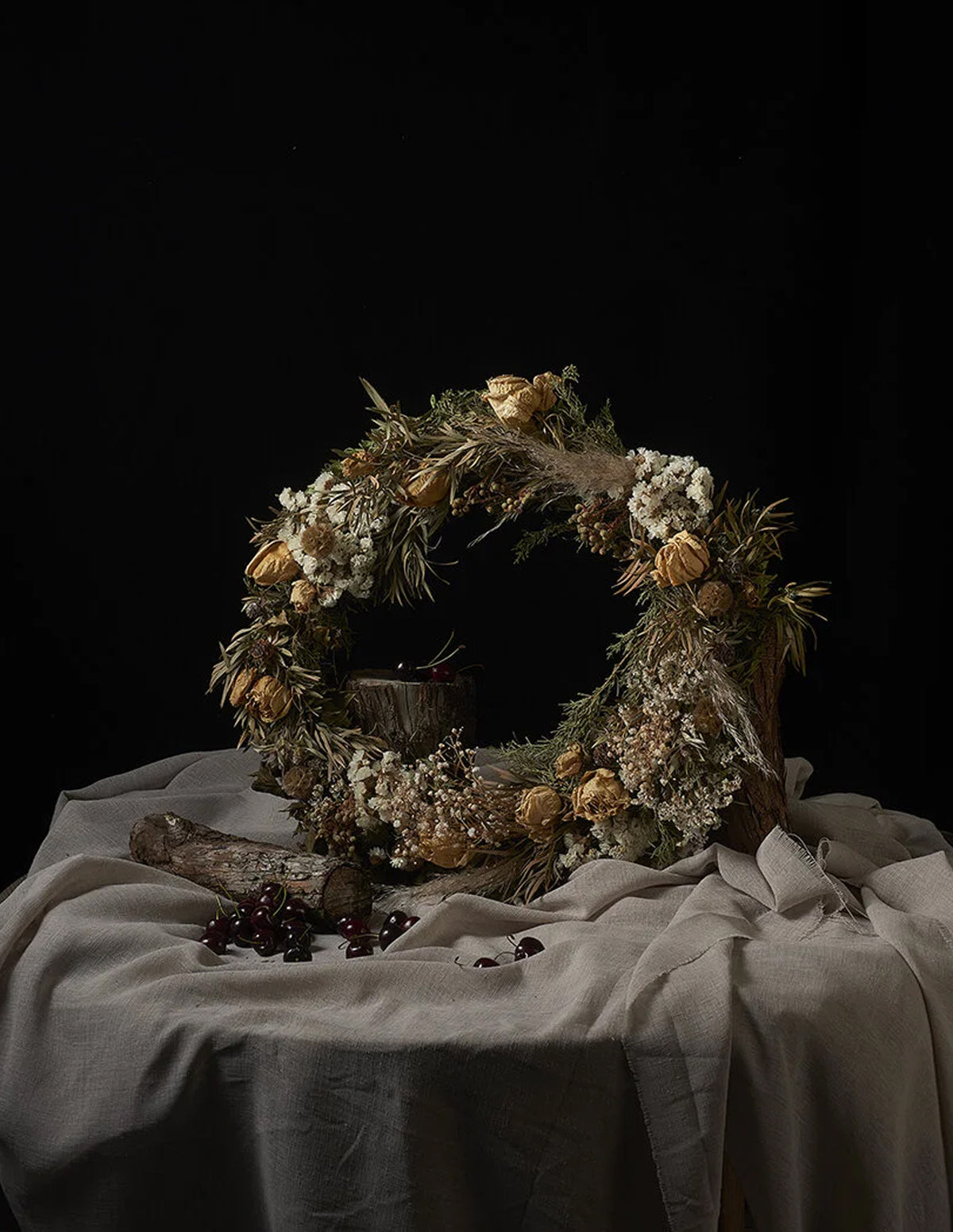 A rustic dried flower wreath with beige, white, and brown botanical elements, displayed on a draped beige fabric-covered table against a solid black background.
