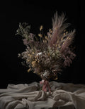 A rustic dried flower bouquet featuring dusty pink pampas grass, beige bunny tails, and white baby's breath, tied with a dusty pink ribbon and set against a draped beige fabric on a solid black background.