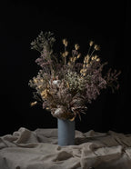 A dried flower arrangement in a textured light blue-gray ceramic vase, set against a solid black background with draped beige linen fabric on the surface.