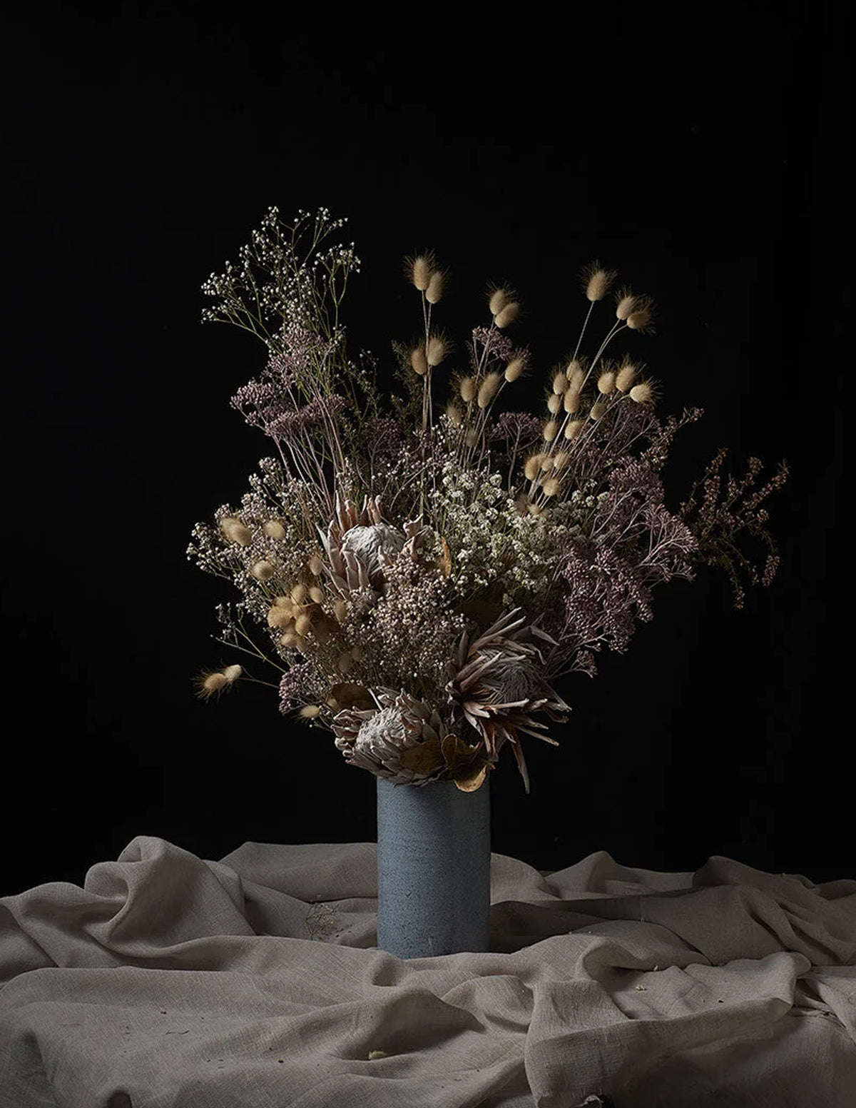 A dried flower arrangement in a textured light blue-gray ceramic vase, set against a solid black background with draped beige linen fabric on the surface.