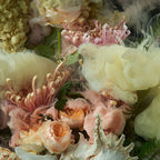 A close-up arrangement of soft pastel flowers and feathery botanical elements, including blush pink roses, pale peach blossoms, cream feathery petals, and textured pale yellow flowers with metallic gold centers.