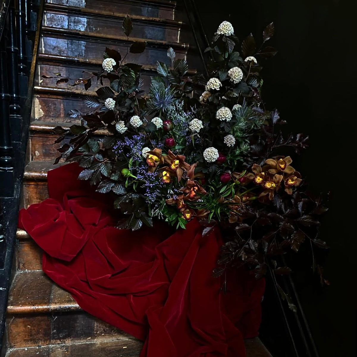 A large, dramatic floral arrangement with dark foliage, white round blooms, purple and yellow accents, and a flowing deep red fabric drape, displayed on a dark wooden staircase.