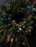 A dark, moody dried floral wreath featuring blue hydrangeas, green pinecones, evergreen branches, and muted purple and brown botanicals against a black background.