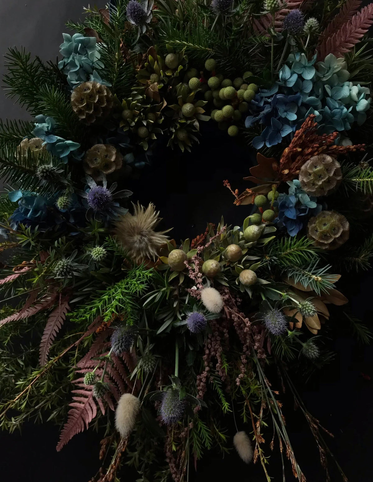 A dark, moody dried floral wreath featuring blue hydrangeas, green pinecones, evergreen branches, and muted purple and brown botanicals against a black background.