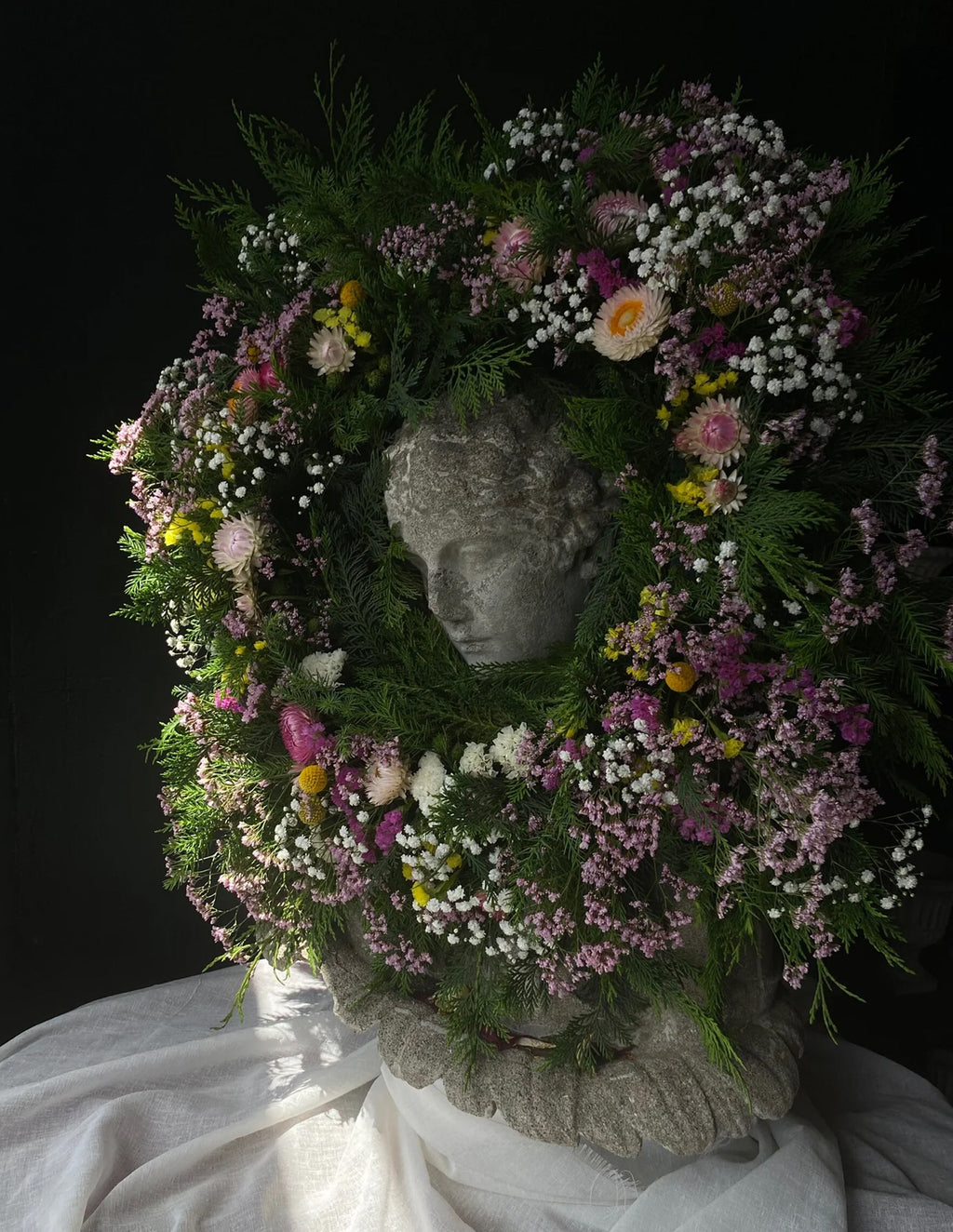 A decorative floral wreath with mixed pink, white, and yellow flowers and green foliage, arranged around a stone classical bust head, set against a dark background with draped white fabric below.