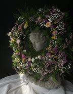 A decorative floral wreath with mixed pink, white, and yellow flowers and green foliage, arranged around a stone classical bust head, set against a dark background with draped white fabric below.