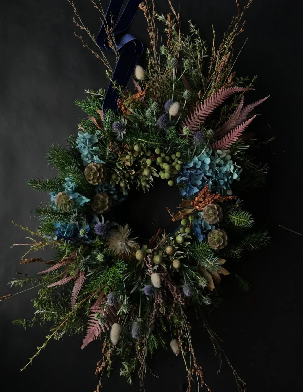 A rustic dried flower wreath featuring blue hydrangeas, pinkish-brown ferns, green thistles, pinecones, and a dark navy blue ribbon at the top, set against a dark background.