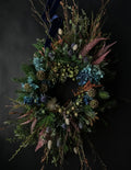 A rustic dried flower wreath featuring blue hydrangeas, pinkish-brown ferns, green thistles, pinecones, and a dark navy blue ribbon at the top, set against a dark background.