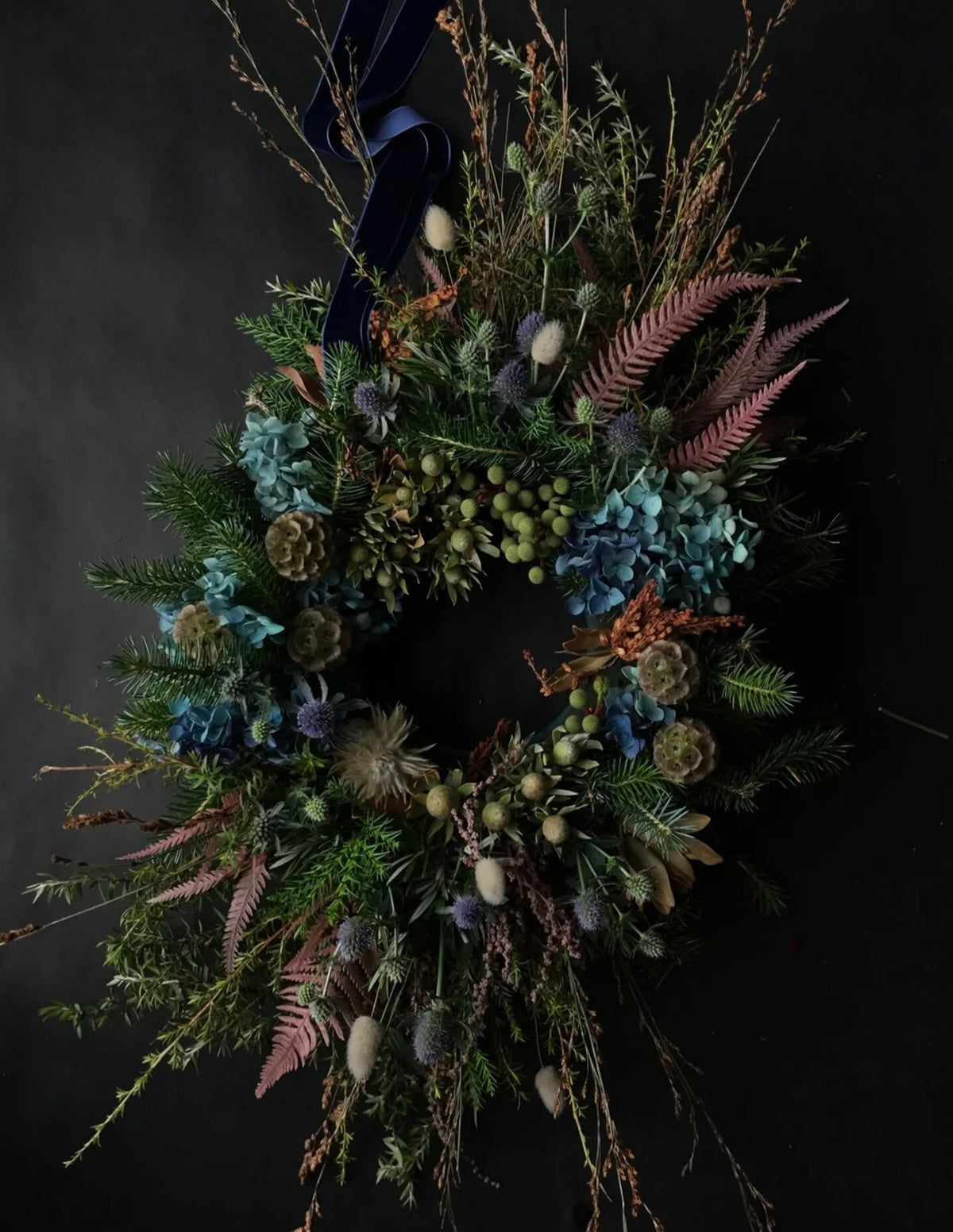 A rustic dried flower wreath featuring blue hydrangeas, pinkish-brown ferns, green thistles, pinecones, and a dark navy blue ribbon at the top, set against a dark background.