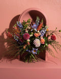 A rustic dried flower arrangement in a terracotta-colored arch-shaped vase, set against a textured beige wall. The bouquet features a mix of purple, white, pink, and brown wildflowers with green stems, casting a soft shadow on the wall.