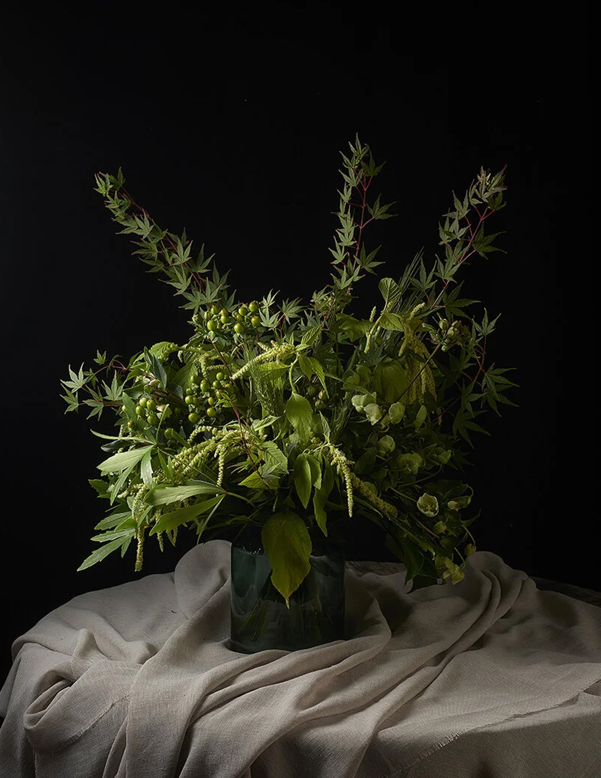 A lush green floral arrangement featuring maple leaves, small green berries, and delicate grasses, presented in a dark glass vase draped over a beige fabric surface against a solid black background.
