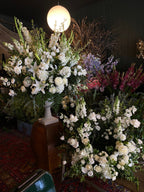Large floral arrangements featuring white lilies, white roses, and green foliage, displayed on a wooden pedestal in an indoor setting with a hanging light visible on the left.