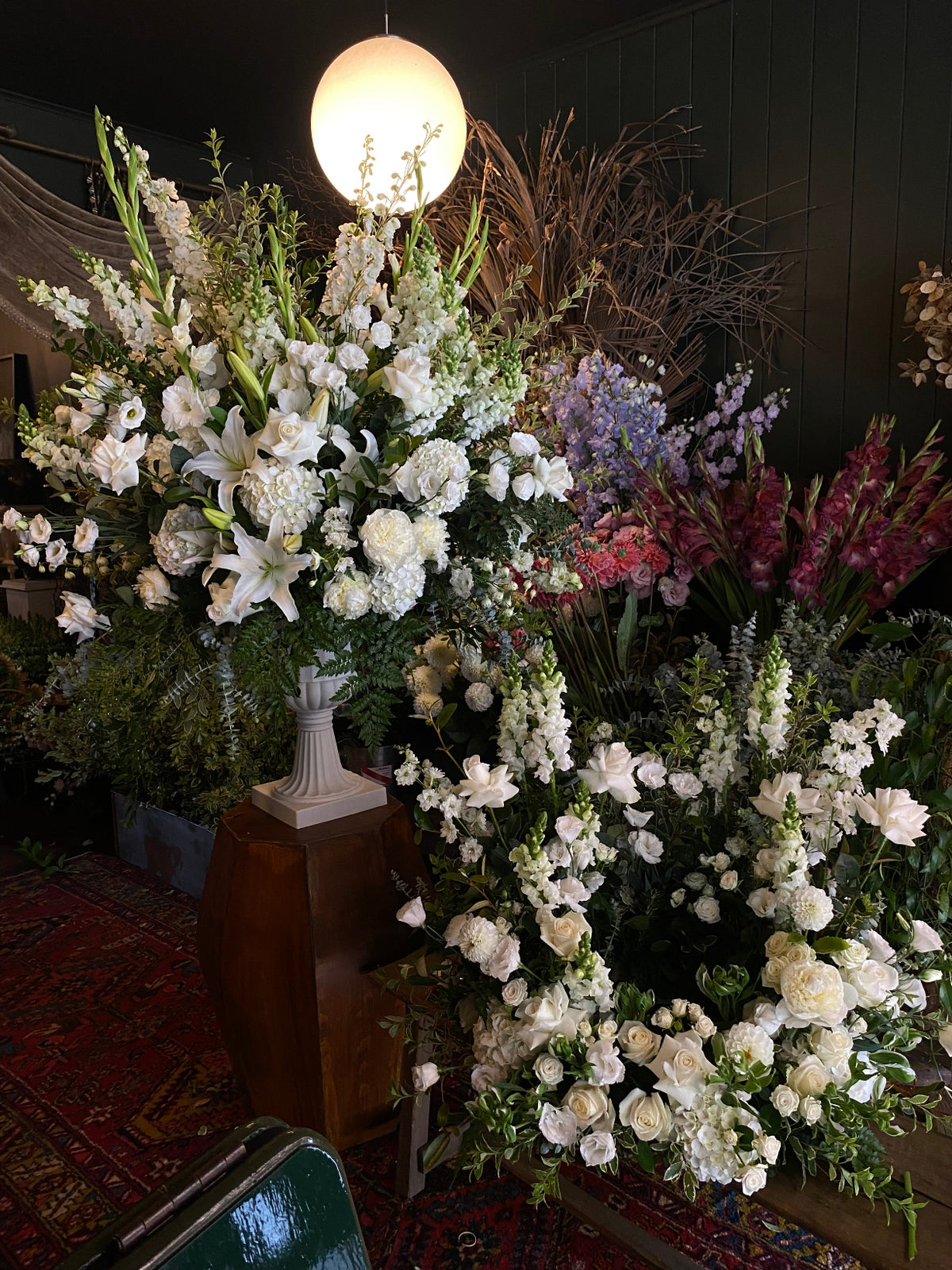 Large floral arrangements featuring white lilies, white roses, and green foliage, displayed on a wooden pedestal in an indoor setting with a hanging light visible on the left.
