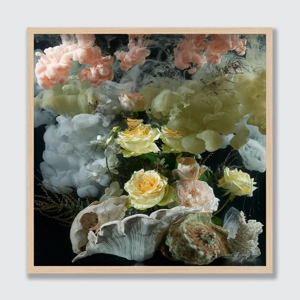 A framed square art print featuring a surreal floral still life with pale pink and yellow roses, white and green smoke-like forms, and large seashells on a dark background, set in a light wood frame.