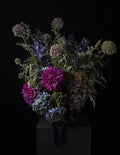 A dark, moody floral arrangement featuring vibrant magenta and lavender flowers, blue hydrangeas, and silvery foliage, displayed on a dark pedestal against a solid black background.