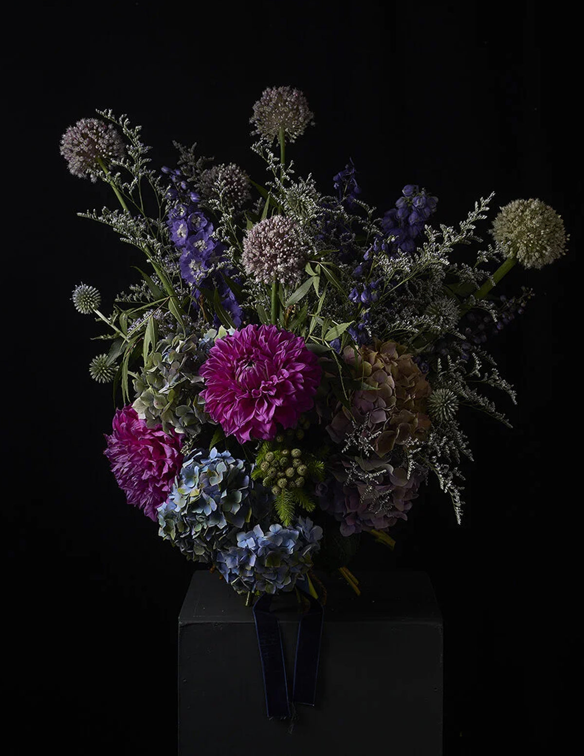 A dark, moody floral arrangement featuring vibrant magenta and lavender flowers, blue hydrangeas, and silvery foliage, displayed on a dark pedestal against a solid black background.