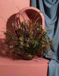 A dried floral arrangement in a textured ceramic pot, styled on a dusty pink pedestal with a draped blue fabric backdrop and a pink arched mirror behind it.