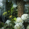 A close-up floral arrangement featuring large white peonies, green leaves, small white flowers, and a textured brownish-purple flower spike against a dark, moody background with water-like drizzle.
