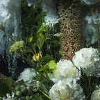 A close-up floral arrangement featuring large white peonies, green leaves, small white flowers, and a textured brownish-purple flower spike against a dark, moody background with water-like drizzle.