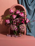 A floral arrangement with pink roses, mauve hydrangeas, and green foliage, styled in front of a beige arched frame and draped blue-gray fabric on a matching beige surface.