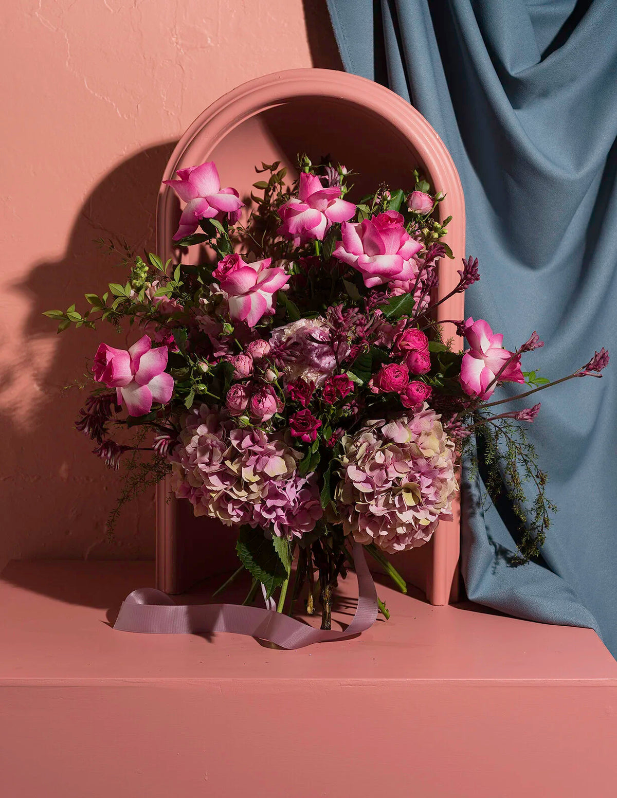 A floral arrangement with pink roses, mauve hydrangeas, and green foliage, styled in front of a beige arched frame and draped blue-gray fabric on a matching beige surface.