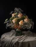 A floral arrangement of peach roses, baby's breath, and green foliage in a dark glass vase, set on draped beige fabric against a black background.
