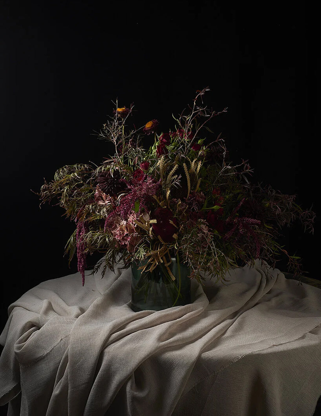 A dried floral arrangement in a dark glass vase, set on a draped beige fabric surface against a solid black background.