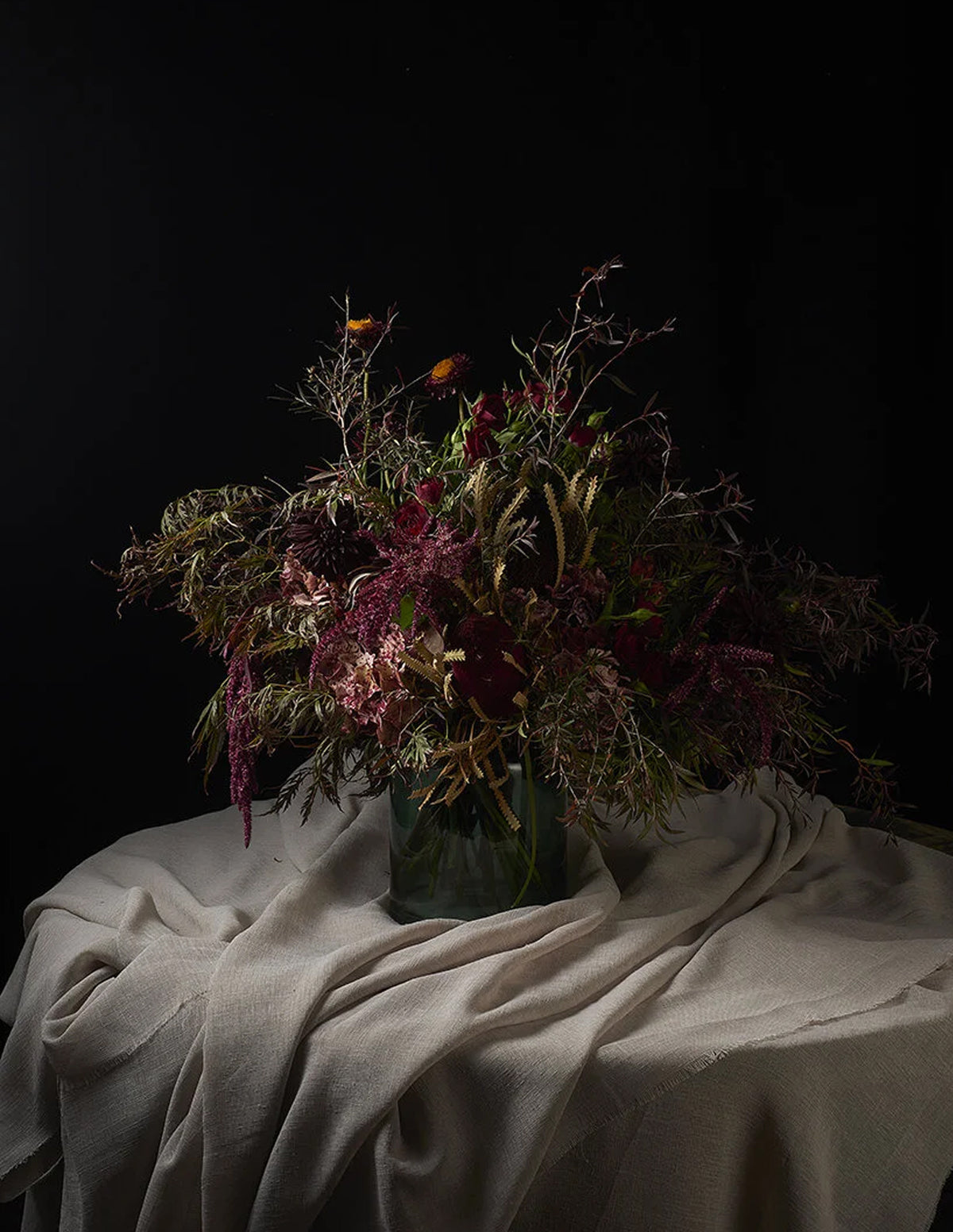A dried floral arrangement in a dark glass vase, set on a draped beige fabric surface against a solid black background.