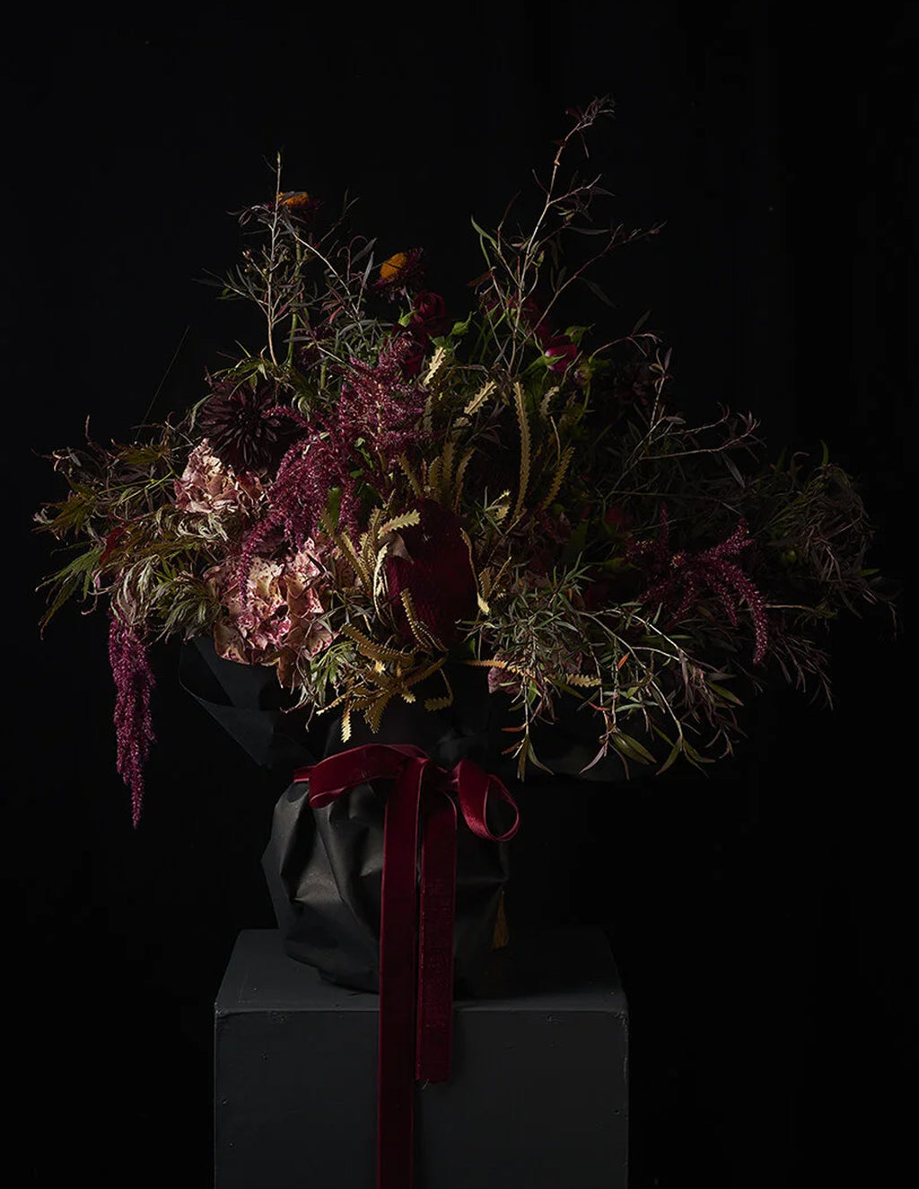 A dark, moody floral arrangement featuring deep burgundy, dusty pink, and olive green botanicals, wrapped in black paper with a prominent burgundy ribbon, displayed on a dark pedestal against a black background.