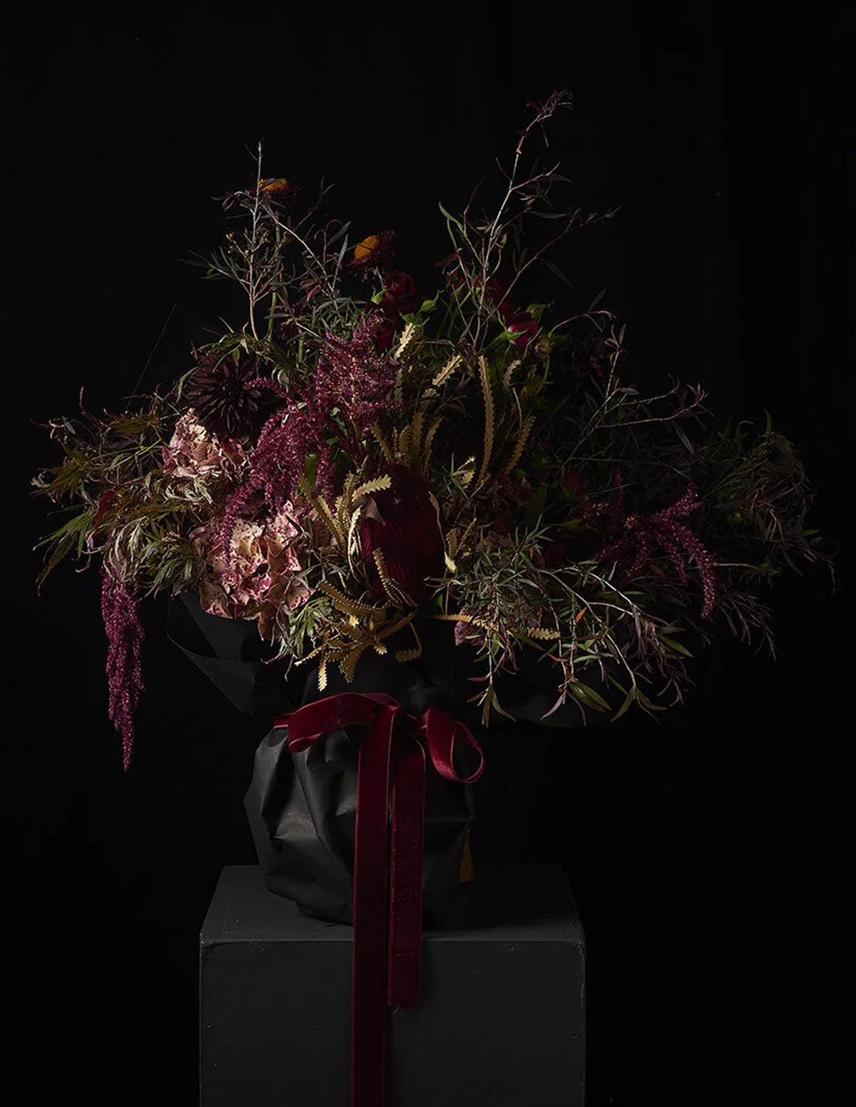 A dark, moody floral arrangement featuring deep burgundy, dusty pink, and olive green botanicals, wrapped in black paper with a prominent burgundy ribbon, displayed on a dark pedestal against a black background.