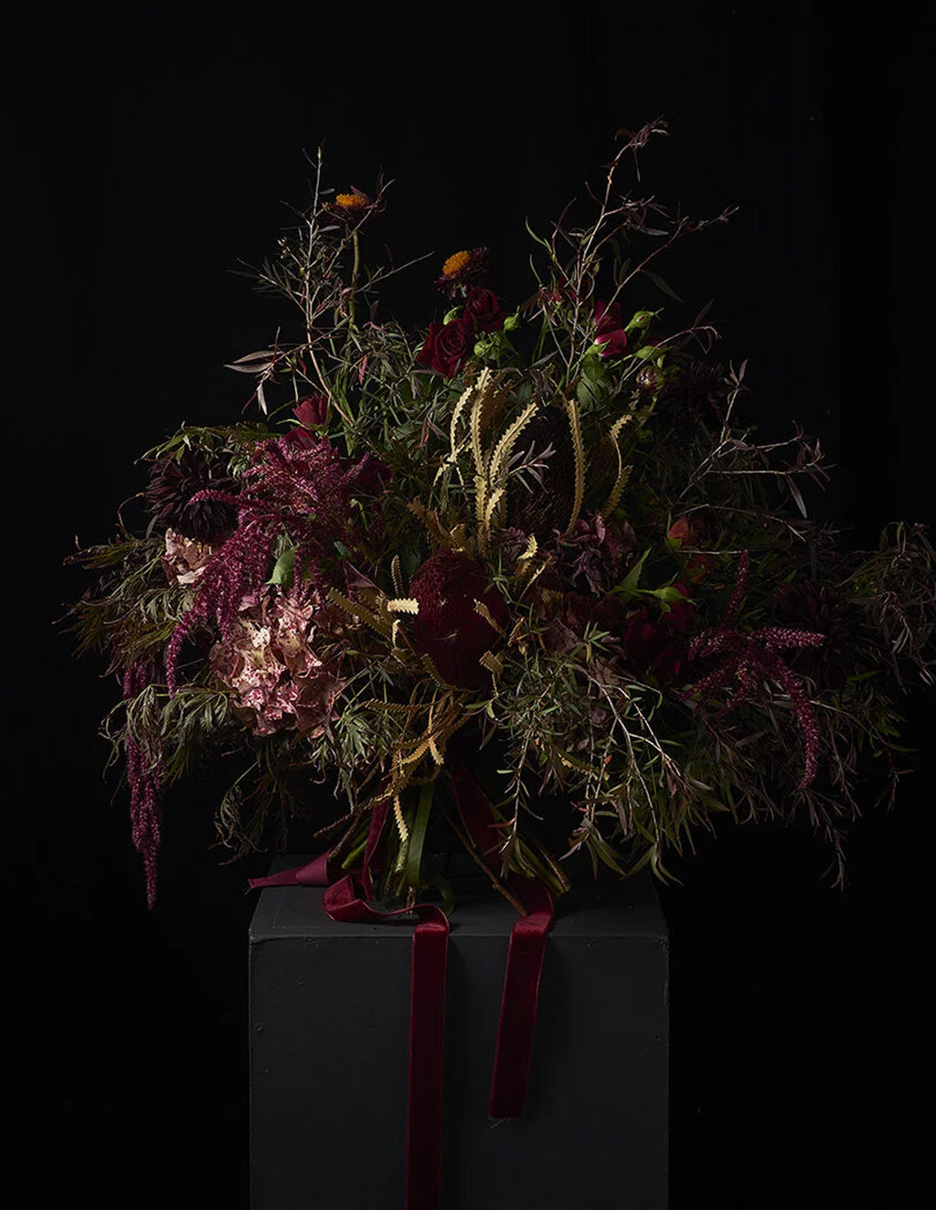 A dark, moody floral arrangement featuring deep burgundy, green, and muted gold botanicals tied with a burgundy ribbon, presented on a dark pedestal against a solid black background.