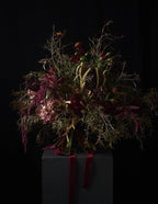 A dark, moody floral arrangement featuring deep burgundy, green, and muted gold botanicals tied with a burgundy ribbon, presented on a dark pedestal against a solid black background.