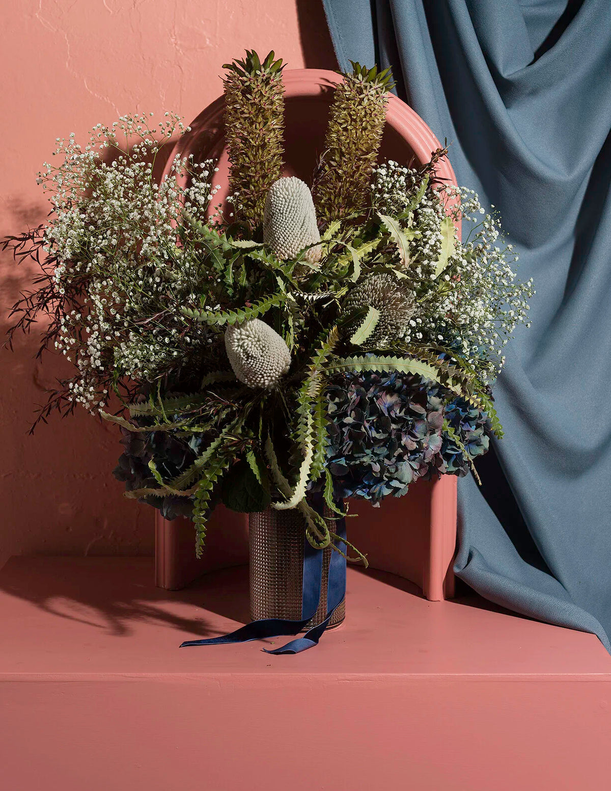 A styled floral arrangement in a textured gray vase, featuring green ferns, white baby's breath, blue hydrangeas, and brown reeds, set against a warm beige wall and draped blue-gray fabric.