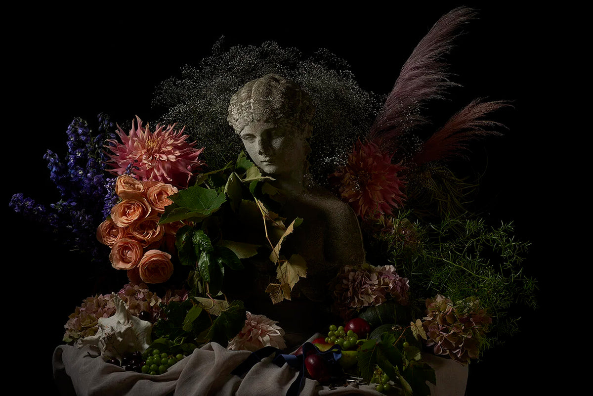 A moody still life arrangement featuring a classical-style stone statue of a woman's head and shoulders, surrounded by an abundant bouquet of flowers including pink dahlias, peach roses, purple delphiniums, green foliage, and feathery dried stems against a solid black background.