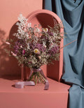 A dried flower bouquet with purple, white, and beige botanicals arranged in a clear glass vase, tied with a mauve ribbon, set on a terracotta pedestal in front of a matching arch and draped blue fabric.